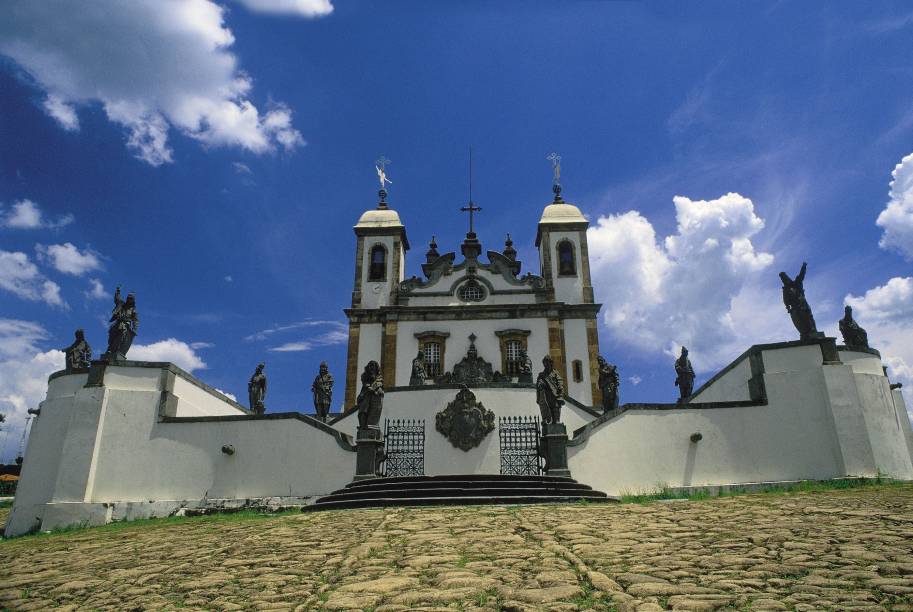 <strong>10. Basílica Bom Jesus de Matosinhos </strong><strong>–</strong><strong> Congonhas</strong> Doente após anos trabalhando em minas de ouro, o imigrante português Feliciano Mendes fez uma promessa: se recuperasse a saúde, mandaria erguer um templo em honra a Bom Jesus de Matosinhos. E assim foi. A obra começou em 1757, mas Mendes morreu oito anos depois, sem saber que de sua iniciativa surgiria um dos maiores tesouros da arte barroca, alçado a Patrimônio Mundial pela Unesco em 1985. A visita à Basílica Bom Jesus de Matosinhos, em Congonhas, se divide em três etapas. À frente, em um plano inclinado com calçamento de pedra, estão as seis Capelas dos Passos, onde 64 imagens de cedro compõem sete cenas da Paixão de Cristo. Algumas foram pintadas por Mestre Athaíde, e parte delas é criação de Aleijadinho. Seu talento atinge o auge na estátua de Daniel, o mais bem-acabado dos 12 profetas em pedra-sabão que enfeitam o adro – os outros são Isaías, Baruc, Jeremias, Ezequiel, Oséias, Joel, Amós, Abdias, Jonas, Naum e Habacuc. Esculpidos entre 1800 e 1805 por Aleijadinho, os profetas formam o destaque do conjunto. Por fim, a igreja exibe traçado inspirado nos santuários portugueses de Matosinhos e Braga. Na parte interna, a igreja abriga relicários também talhados por Aleijadinho, rica decoração rococó e pintura do teto assinada por Mestre Athaíde. Anexa fica a sala de ex-votos, transformada em museu <a href="https://www.booking.com/city/br/congonhas-br.pt-br.html?aid=332455&label=viagemabril-13-atracoes-historicas-mg" target="_blank" rel="noopener"><em>Reserve já o seu hotel em Congonhas (MG)</em></a>