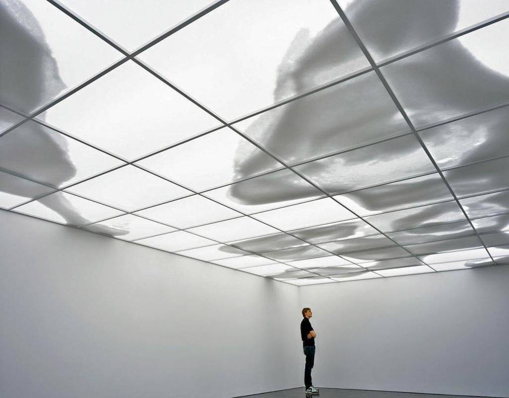 Inhotim, Brasil Uma pessoa, sozinha, se encontra em um quarto desprovido de móveis, com um teto onde sombras se movem, tomando a forma de nuvens