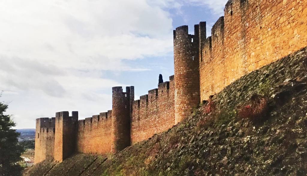 Em cima de uma colina, o muro grosso de uma fortificação medieval se ergue, pontuado por torres de observação.