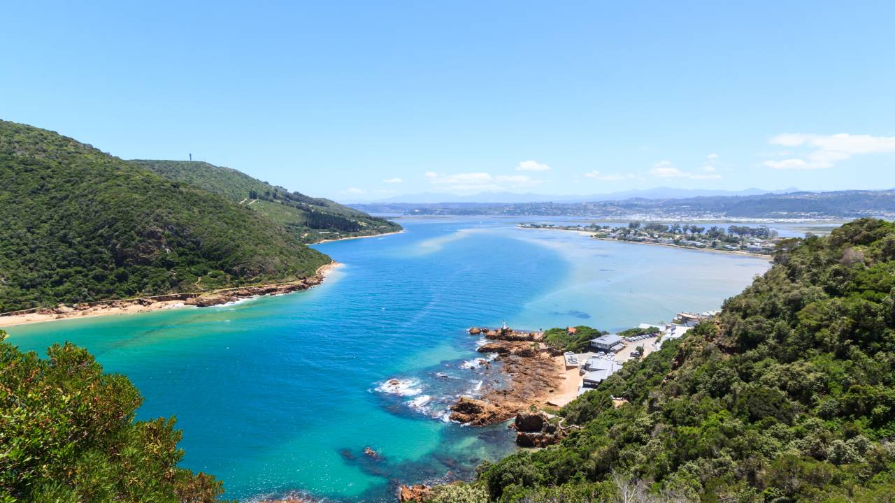 Colinas terminam em faixas de areia de uma lagoa, a qual desagua. No fundo, o contorno de uma cidade. Atrás, fora da foto, está o mar