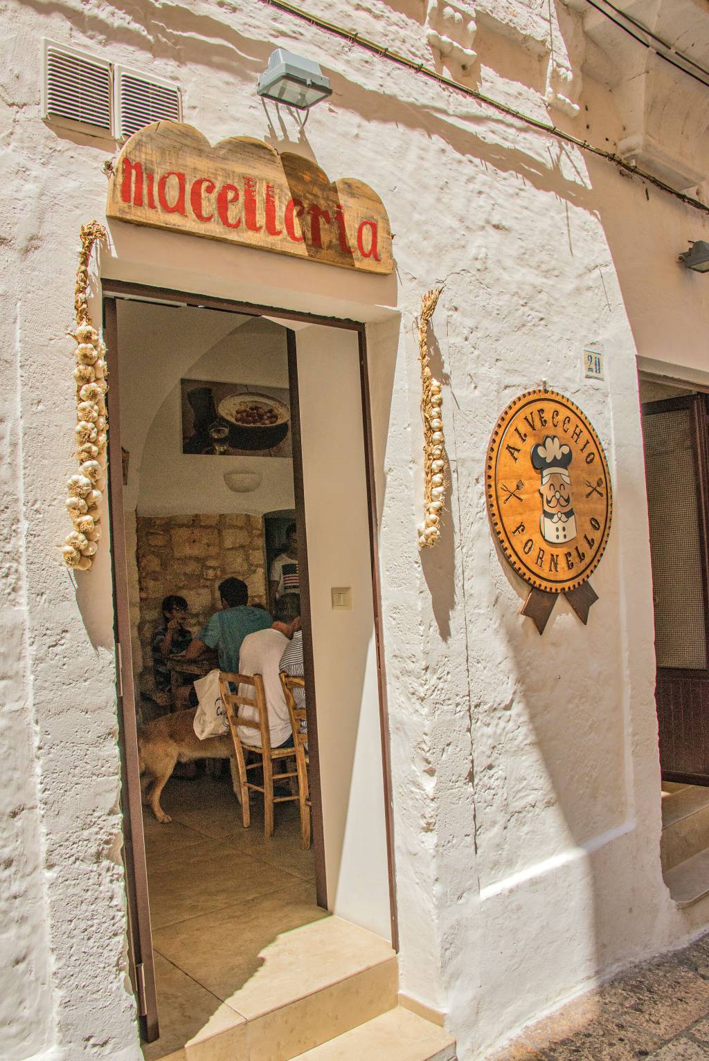 Macelleria Al Vecchio Fornello, em Cistertino, Puglia, Itália Alta parede branca, com uma porta em que os batentes estão decorados com correntes de alho. Dentro, é possível ver várias meses e cadeiras com clientes consumindo alimentos