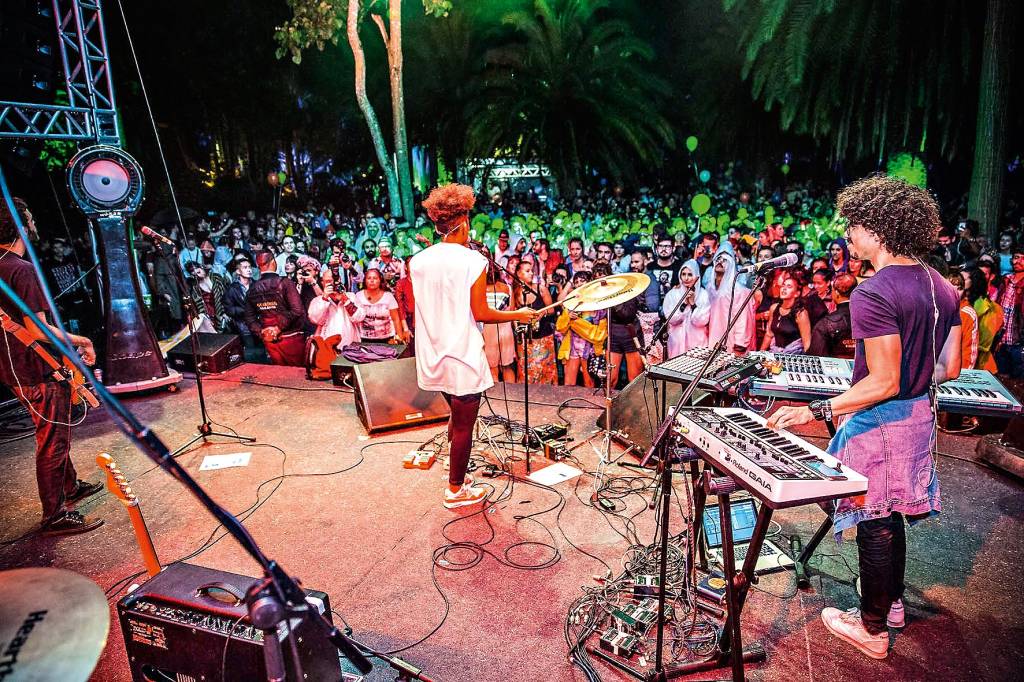 mahmundi, Inhotim, Brasil Vista de cima do palco de um show de música, com uma banda de estilo alternativo e uma multidão de espectadores igualmente vestidos.
