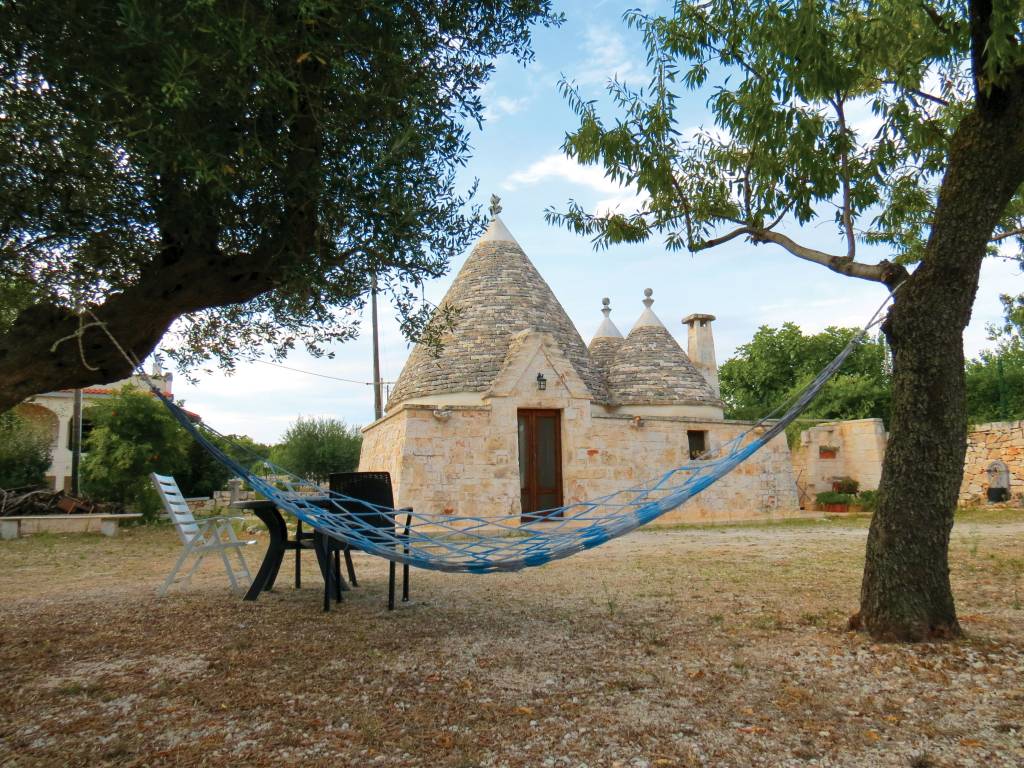 trullo-na-puglia-italia Casa em estilo trullo, com um quintal na frente, no qual duas árvores suportam uma rede