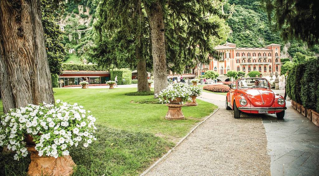 Um fusca conversível percorre o caminho de saída do suntuoso jardim do hotel Villa d'Este, que se encontra ao fundo