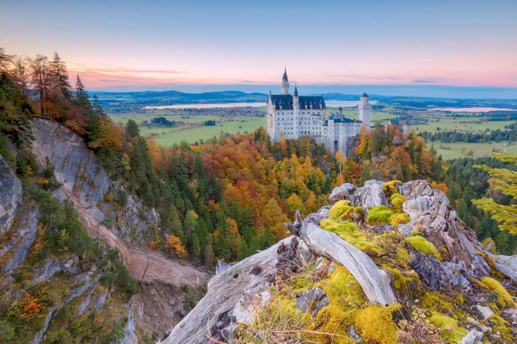 A imagem mostra um castelo romântico sobre as montanhas. Ao seu redor, as folhas atingem tons de vermelho, laranja e amarelo.