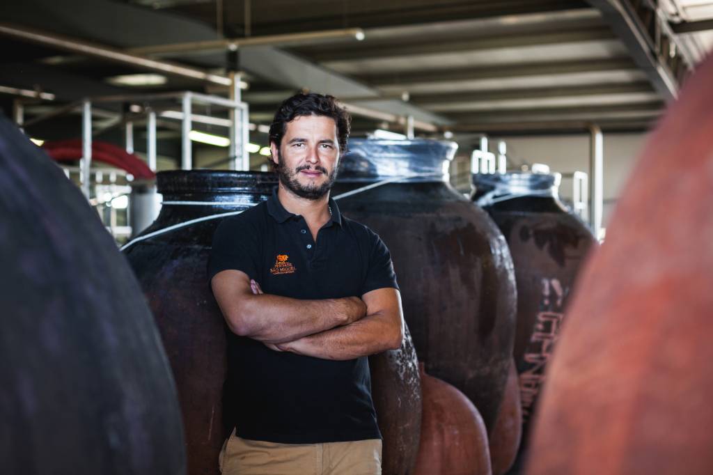Alexandre Relvas 21 A imagem mostra Alexandre Relvas, um homem branco, com cabelo e barba castanhos. Ele está em pé, em frente de barris de madeira de vinho.