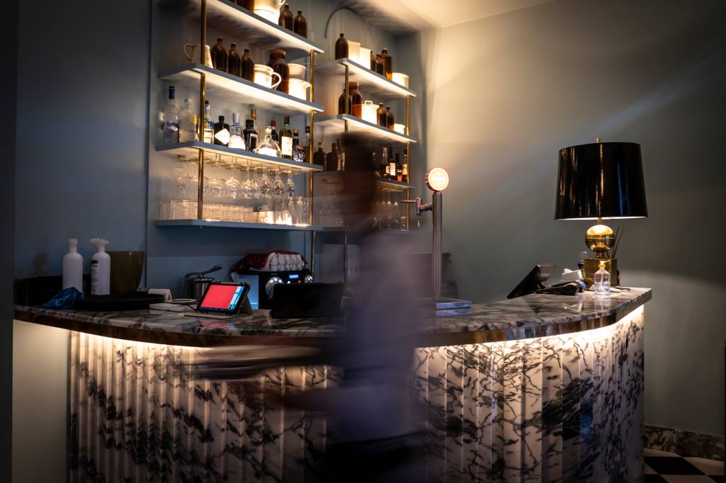 Drogaria1 Bar com um balcão escuro de mármore e uma pessoa passando na frente, desfocada