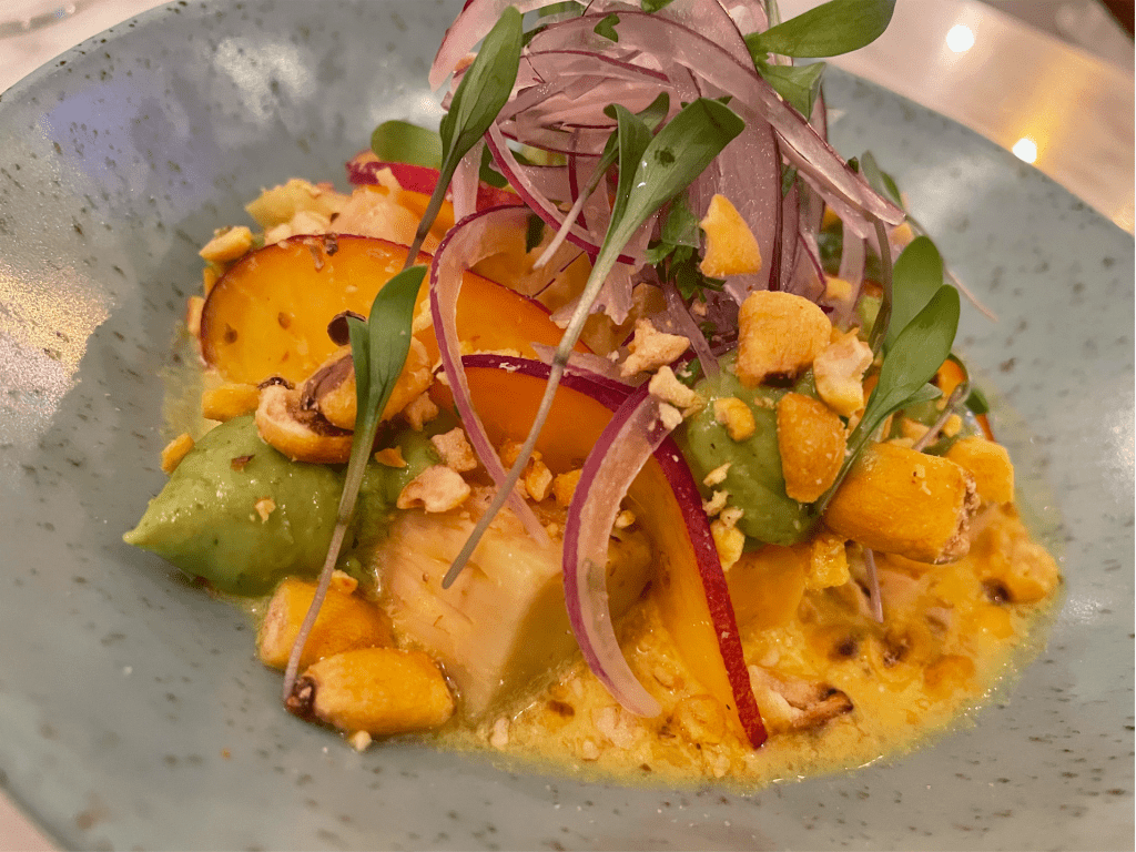 SenhorUva3 Prato de cerâmica cinza com cubos de peixe, fatias de nectarina, cebola roxa, milho frito e um caldo amarelo ao fundo