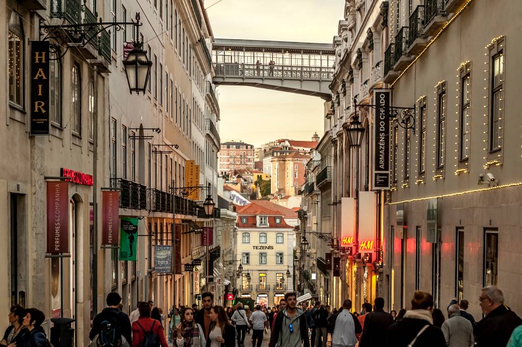 1622-2016_BrunoBarata Rua cheia de pessoas com construções antigas dos dois lados e uma ponte de ferro ligando os dois lados