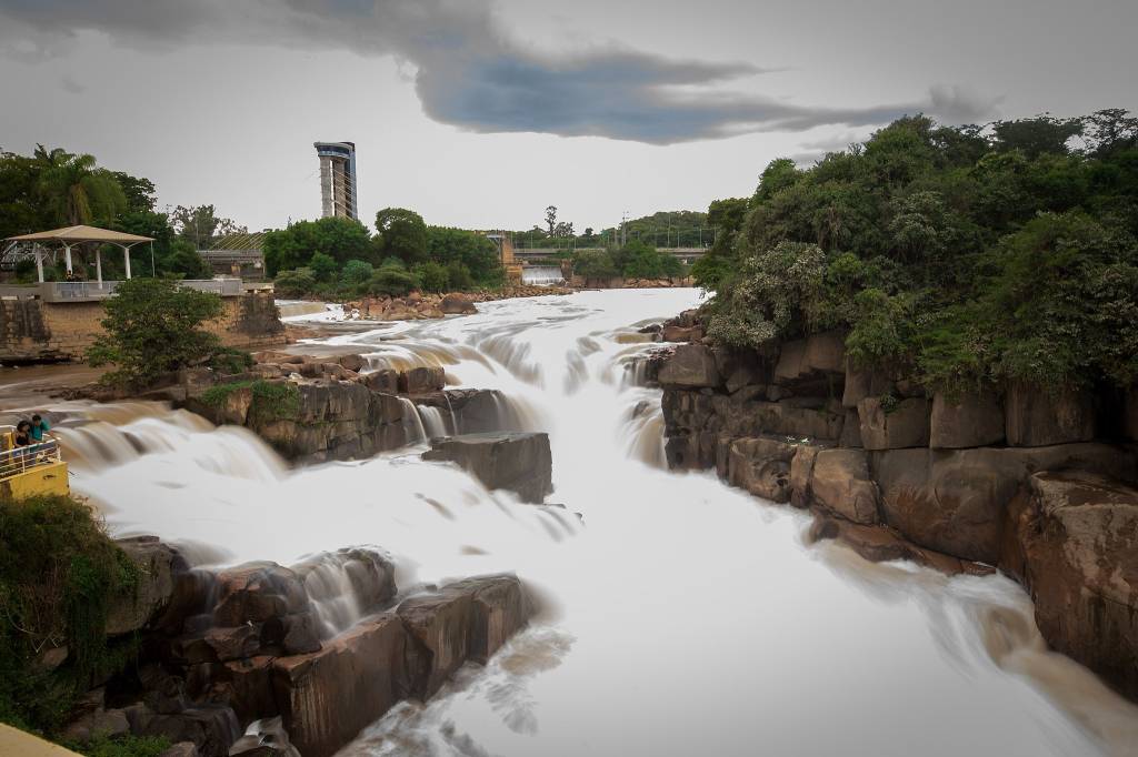 A imagem mostra uma cachoeira no centro. Ao seu redor há vegetação e uma ponte estaiada pode ser vista no fundo da foto.