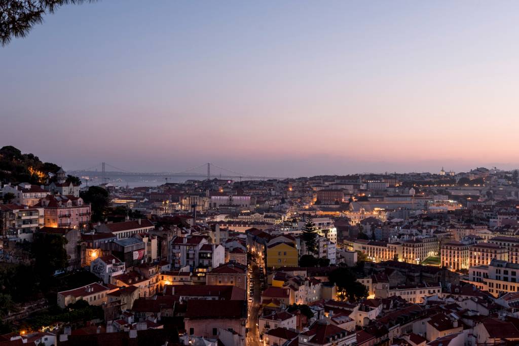 O casario de Lisboa descendo a colina ao anoitecer, com o rio Tejo e a Ponte 25 de Abril ao fundo