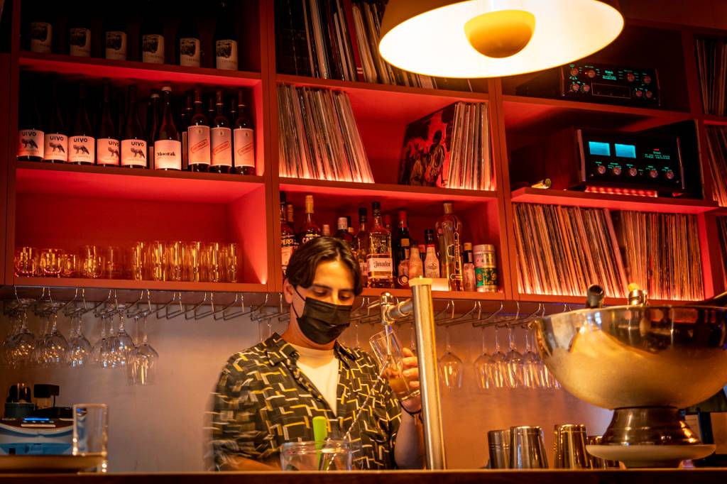 3 Barman servindo cerveja no balcão de um bar, com uma prateleira atrás recheada de copos, garrafas de bebidas e discos de vinil