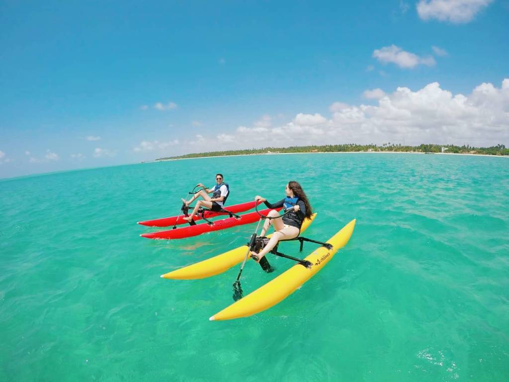 A imagem mostra duas pessoas pedalando uma espécie de bicicleta aquática que possui duas partes de caiaques no lugar das rodas. Eles estão sobre um mar de verde intenso e ao fundo vê-se um trecho de praia e o céu azul com poucas nuvens.