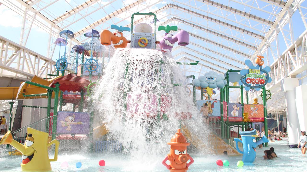 A imagem mostra uma área de piscinas infantis. É possível ver uma grande instalação de brinquedos em formato de torneiras, hidrantes e guarda-chuvas, além de um grande balde que despeja água sobre a rasa piscina.