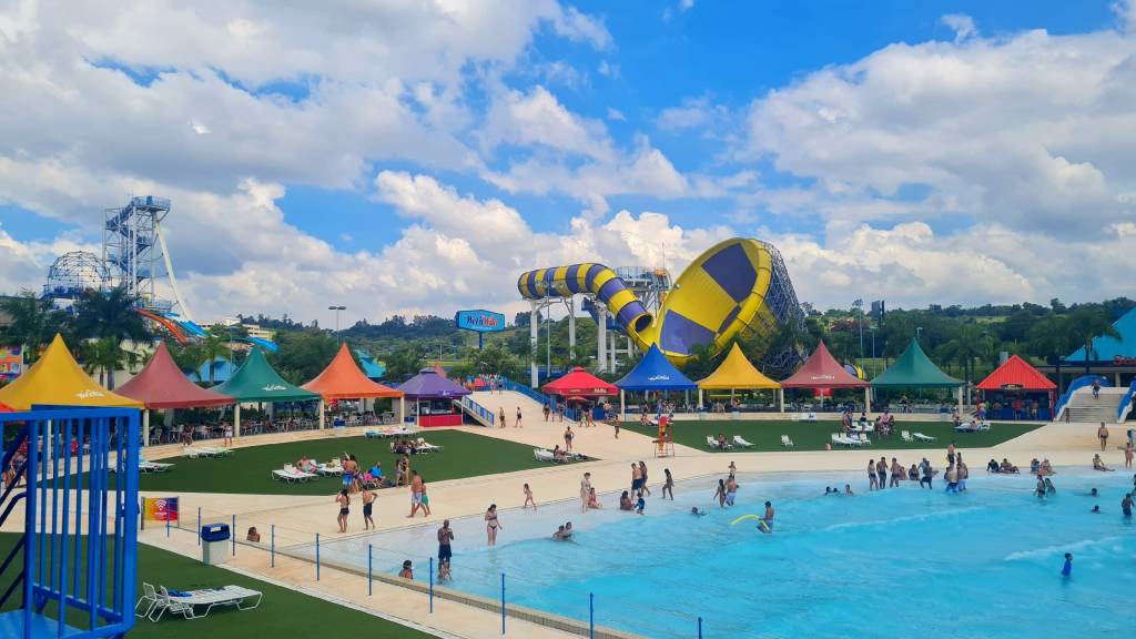 Wet’n Wild – Tendas, ondas e Vortex Na fotografia é possível observar uma piscina de ondas cercada por tendas coloridas. Ao fundo, vê-se um enorme brinquedo que se parece com um funil amarelo e roxo.