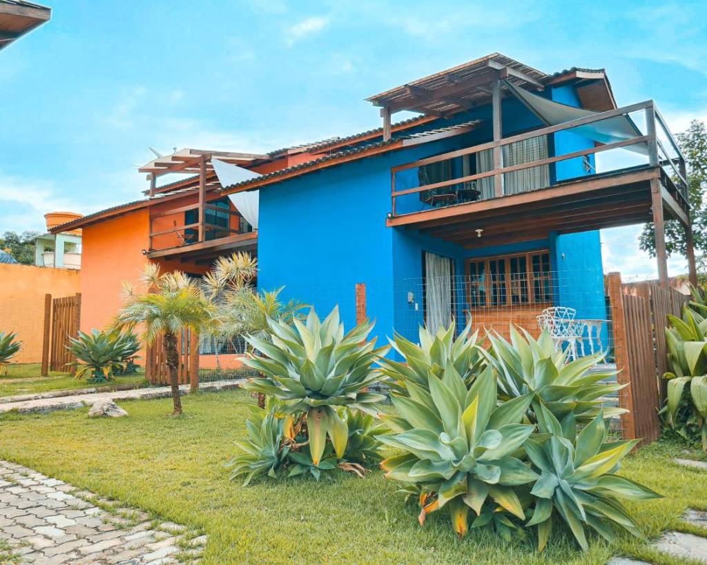 Capim Canoa, Alto Paraíso de Goiás, Goiás A fotografia colorida mostra duas casas de dois andares, sendo uma azul e outra laranja. Elas estão cercadas por grama e arbustos pontiagudos.