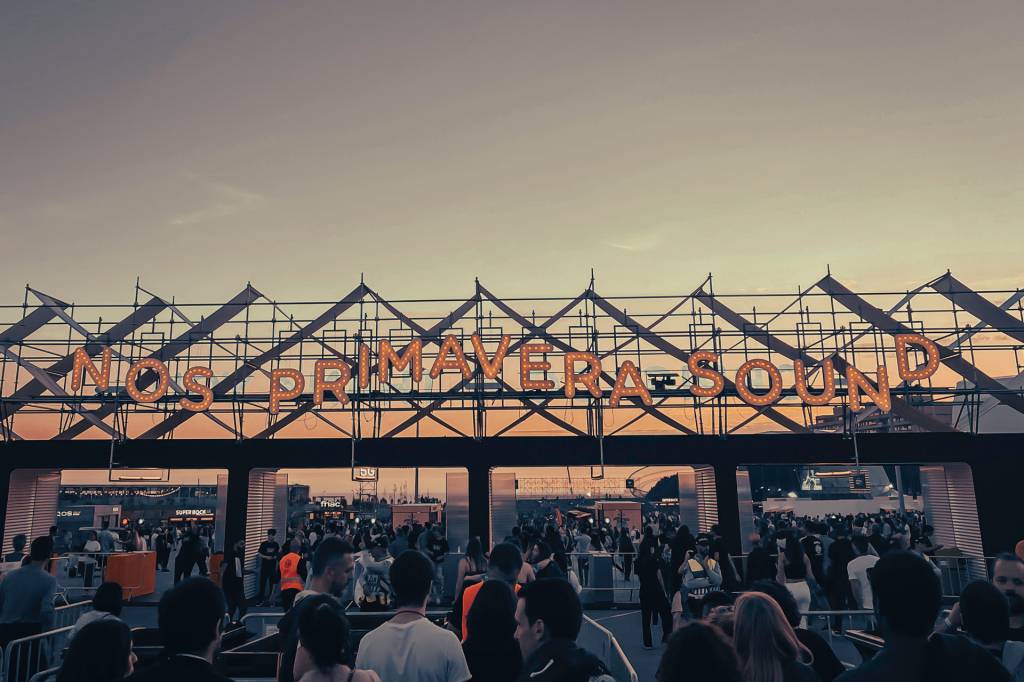 Uma multidão de pessoas em fila para entrar no parque onde aconteceu um festival de música, sob um letreiro luminoso escrito NOS Primavera Sound