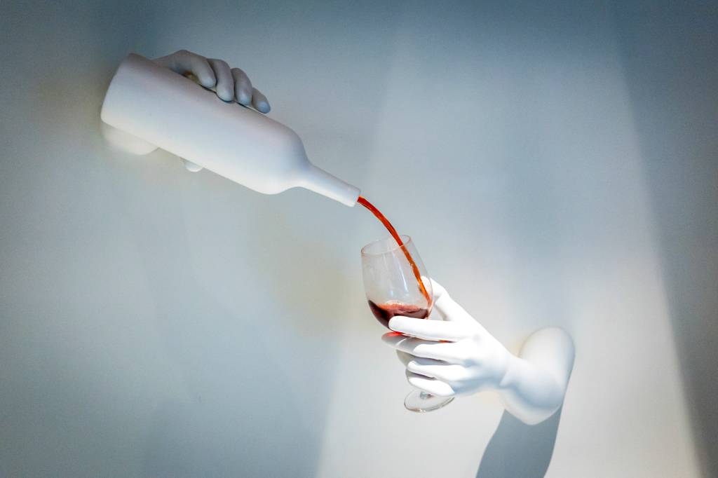 80 Escultura em parede branca mostra uma mão segurando uma garrafa, também brancas, servindo vinho tinto a uma taça segurada por outra mão
