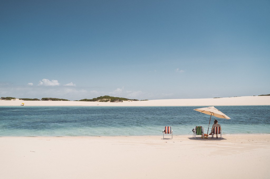 A foto colorida mostra uma pessoa sentada em uma cadeira de praia de frente para uma lagoa em Lençóis Maranhenses