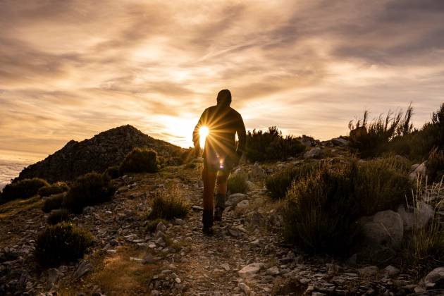 Homem caminhando em uma trilha com o sol a nascer no fundo