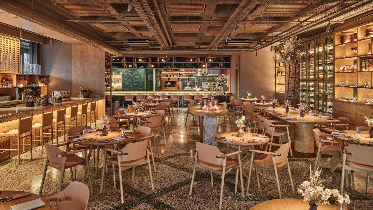 A imagem colorida mostra o salão de um restaurante com muita madeira e cimento.