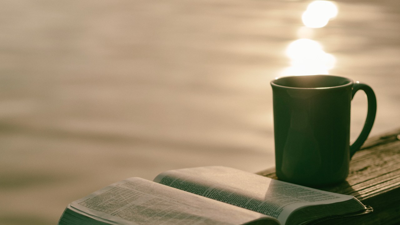 Um livro e uma caneca de café e, no fundo, o mar com o reflexo do sol