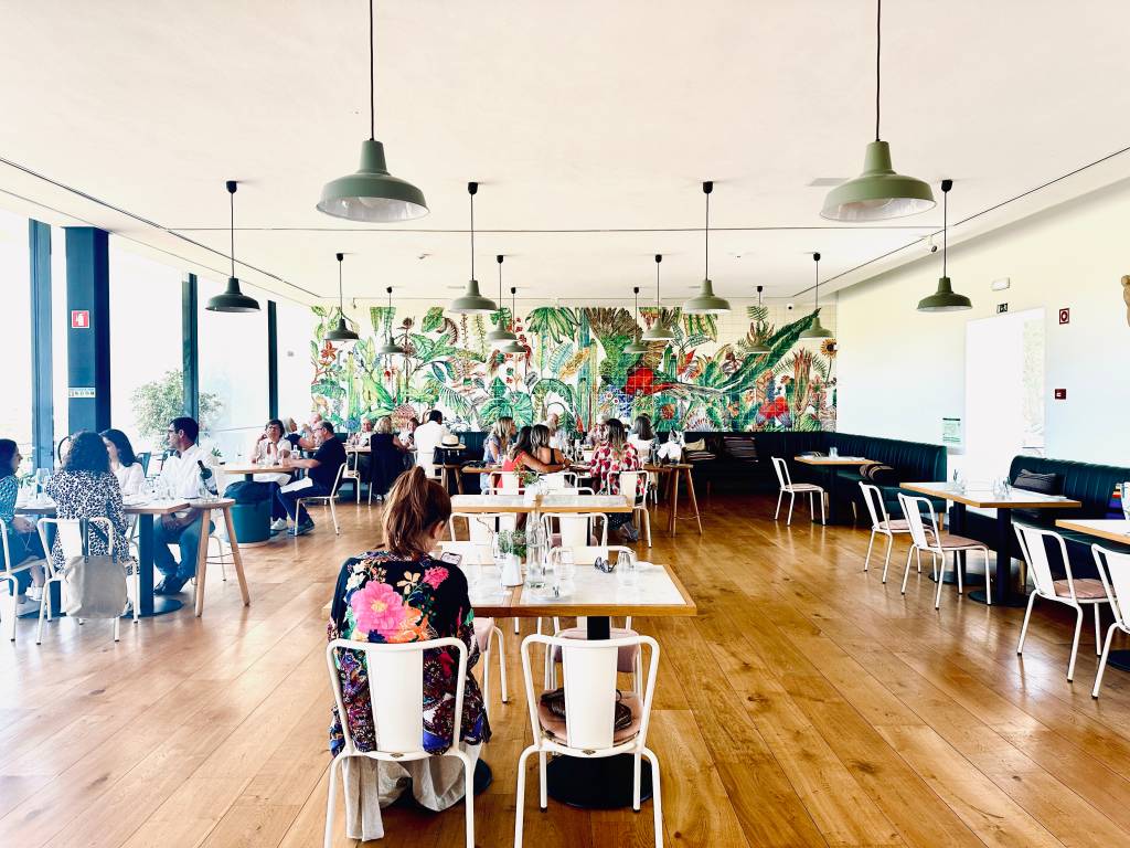 IMG_9521 Sala de um restaurante com piso de madeira, pequenas mesas com cadeiras brancas, luminárias pendendes e um grande painel de azulejos coloridos com desenhos de plantas tropicais ao fundo
