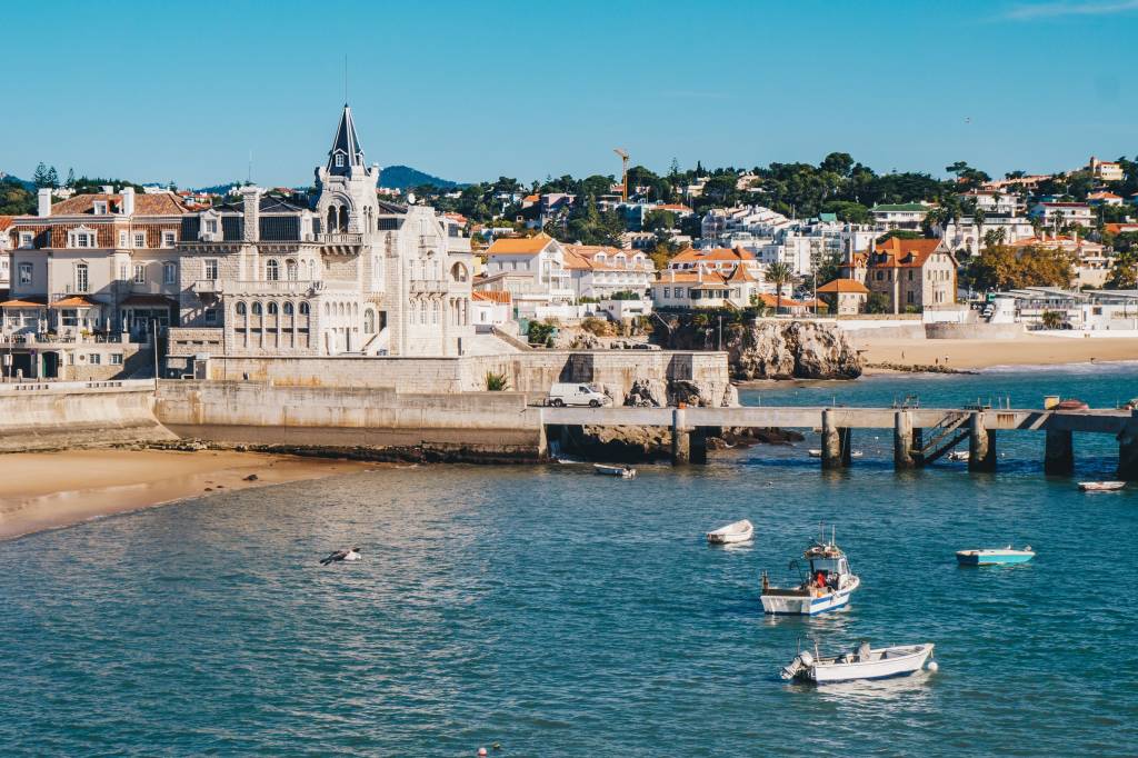 calin-stan-1V-hH2oF3ls-unsplash A foto mostra num primeiro plano o mar azul com barquinhos de pesca e, atrás, casas antigas do centrinho de Cascais