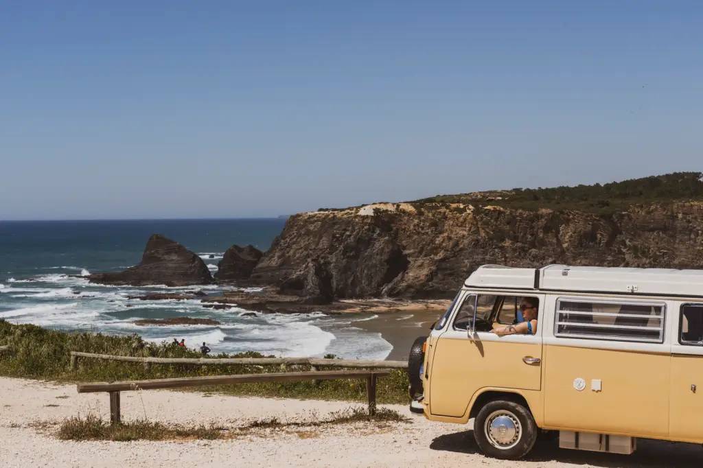 Foto colorida mostra uma praia selvagem com uma kombi antiga em primeiro plano