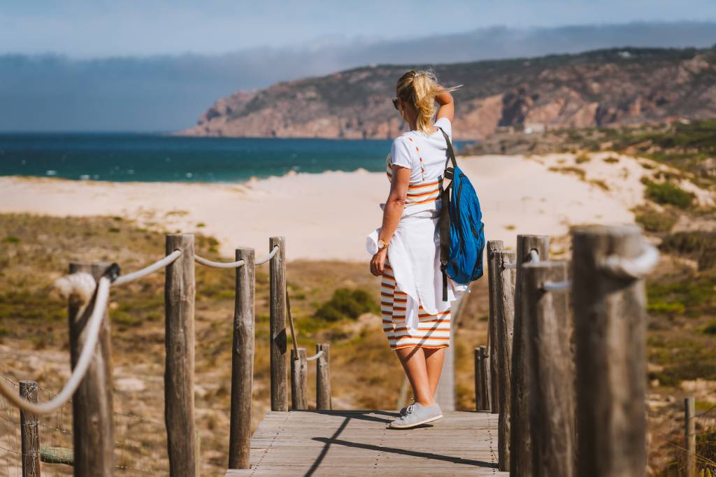 Foto colorida mostra uma mulher a olhar do alto uma praia de areias brancas e águas azuis