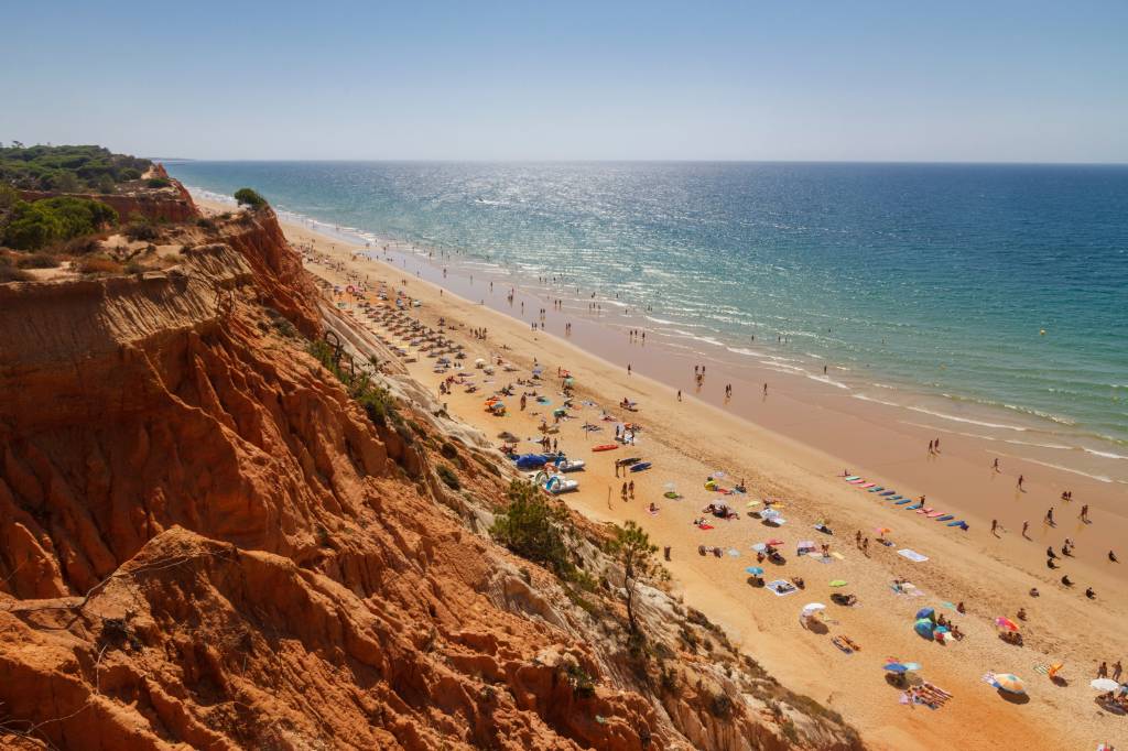 Foto colorida mostra uma praia vista do alto de suas falésias coloridas. Lá embaixo, banhistas e guarda-sóis se espalham na areia