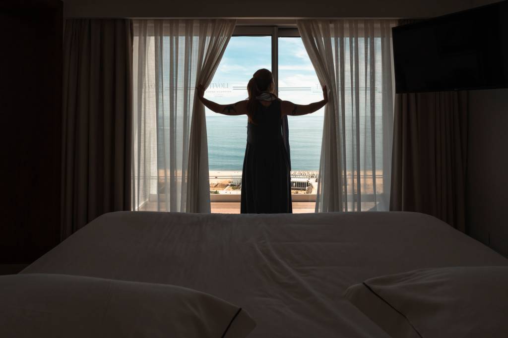 22 Cama do quarto do hotel, com uma mulher abrindo as cortinas, através das quais se vê o mar