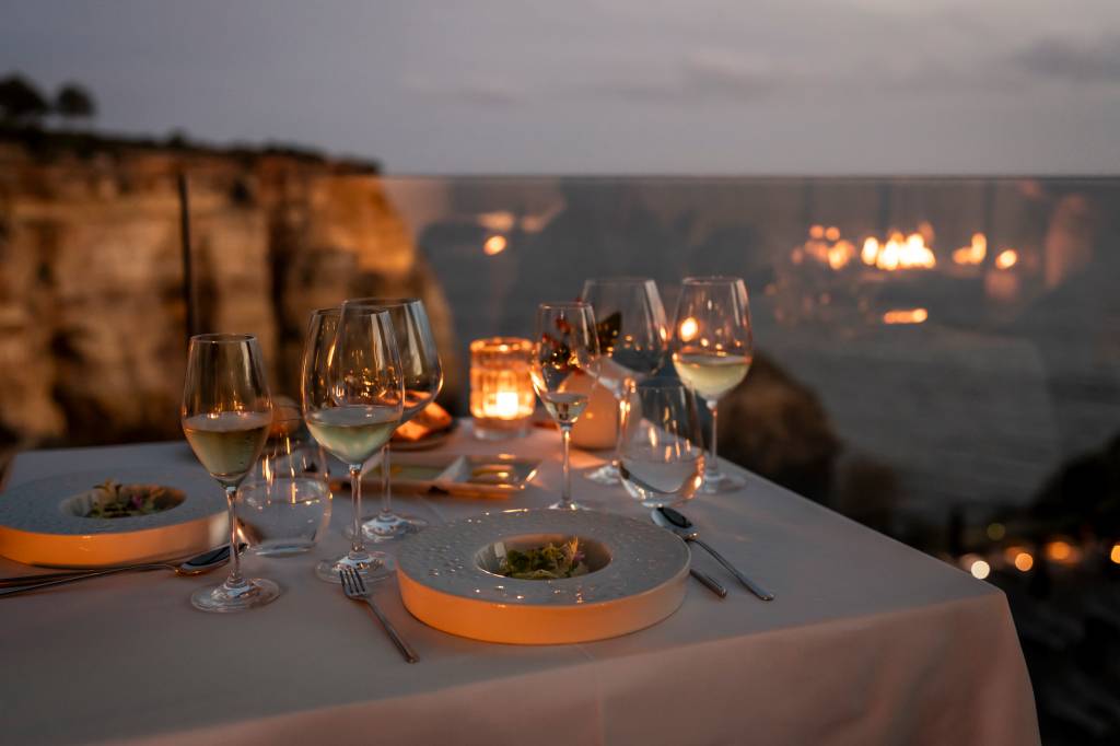 6 Mesa à luz de velas, com taças de vinho e pratos; ao fundo, falésia e mar