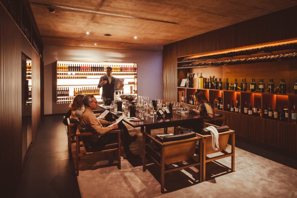 1 2 numa sala cercada de paredes com garrafas, um sommerlier conduz um workshop a três pessoas sentadas numa mesa, onde estão copos e garrafas de vinho