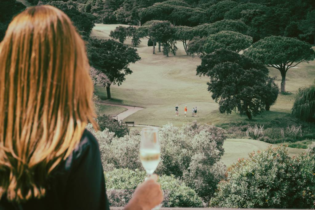 2024_Nuvem-385 Mulher observa um dos campos de golfe do hotel da varanda de sua suíte, com uma taça de espumante na mão