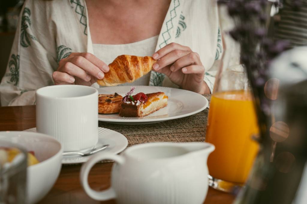 2024_Nuvem-414 Detalhe de uma mesa de café da manhã do hotel: hóspede a segurar um croissant, com um copo de suco de laranja à frente