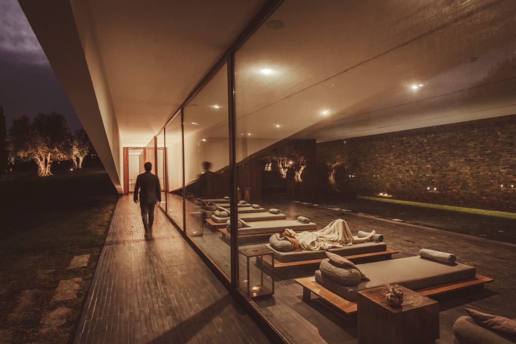 61 2 A imagem mostra o spa do hotel, com uma pessoa a caminhar do lado de fora e, do lado de dentro, espreguiçadeiras com os hóspedes em volta da piscina aquecida