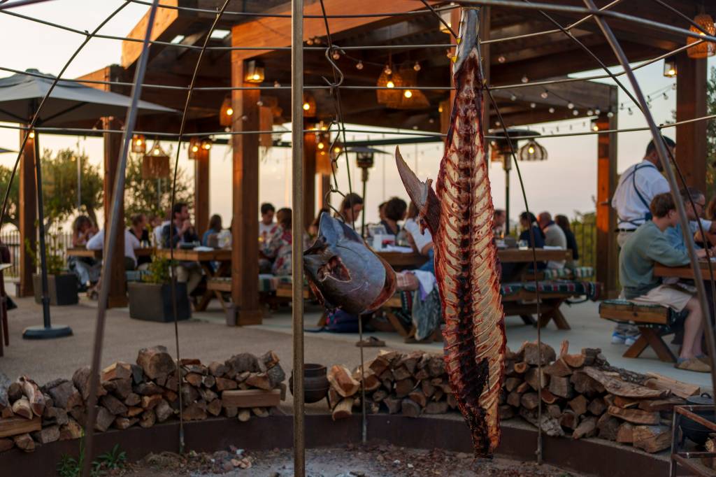 Imagem do restaurante com mesas ao ar livre e pessoas a jantar, e o esqueleto de um enorme atum no fogo de chão
