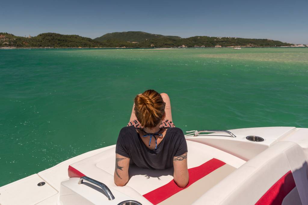 Mulher deitada num barco cercado de aguas verde-esmeralda. Ao fundo vê-se montanhas e praias