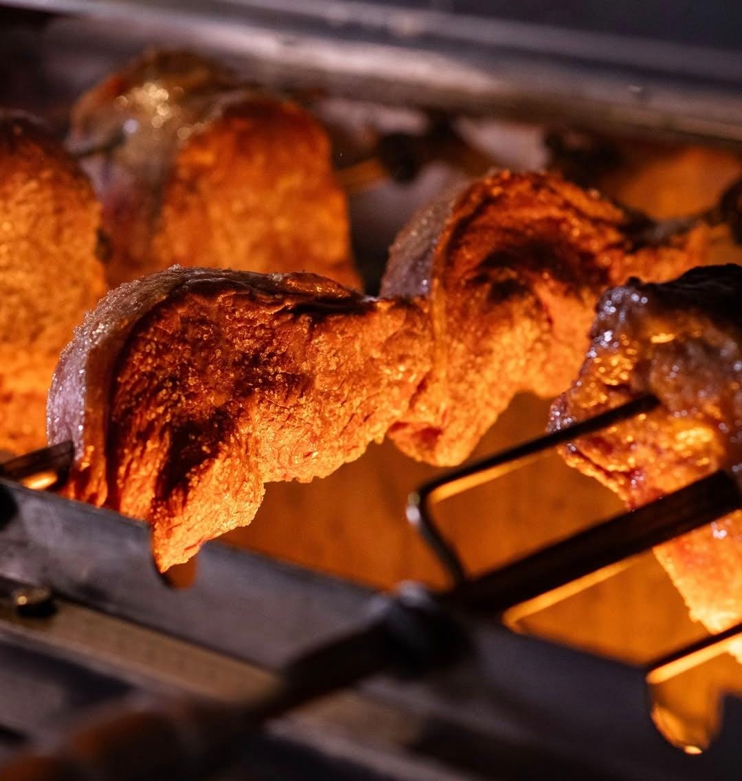 Pedaços de carne bovina, possivelmente picanha, assando em espetos metálicos dentro de uma churrasqueira, com a gordura derretendo e a carne dourada pela brasa