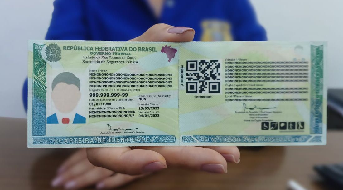 Mão feminina segura a nova Carteira de Identidade Nacional (CIN) com fundo azul. O documento verde e amarelo exibe dados fictícios, QR Code e mapa do Brasil, destacando a modernização da identificação civil