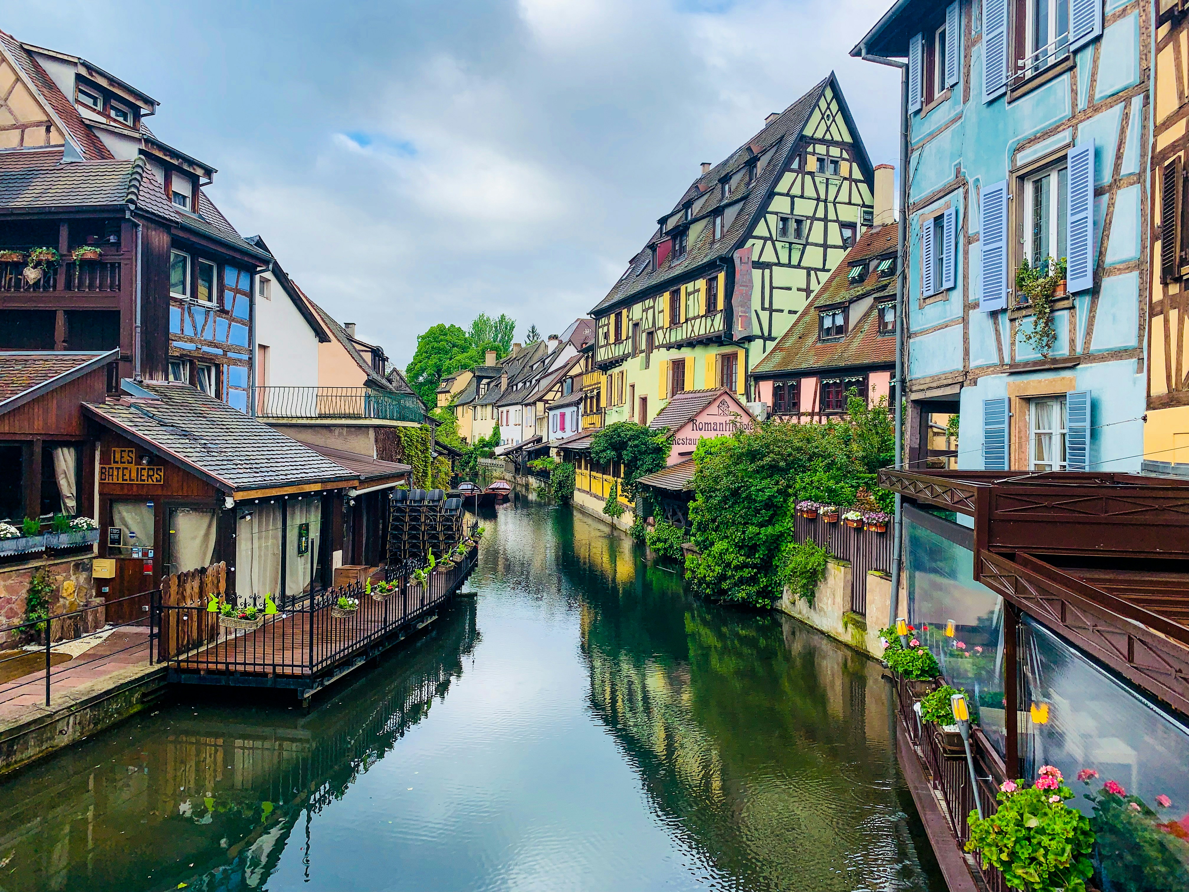 Canal estreito ladeado por casas coloridas de arquitetura tradicional europeia, com telhados inclinados e janelas com venezianas. Há vegetação exuberante e flores em vasos nas varandas e margens, sob um céu parcialmente nublado. Um restaurante com deck de madeira flutua à esquerda, e a água reflete as construções