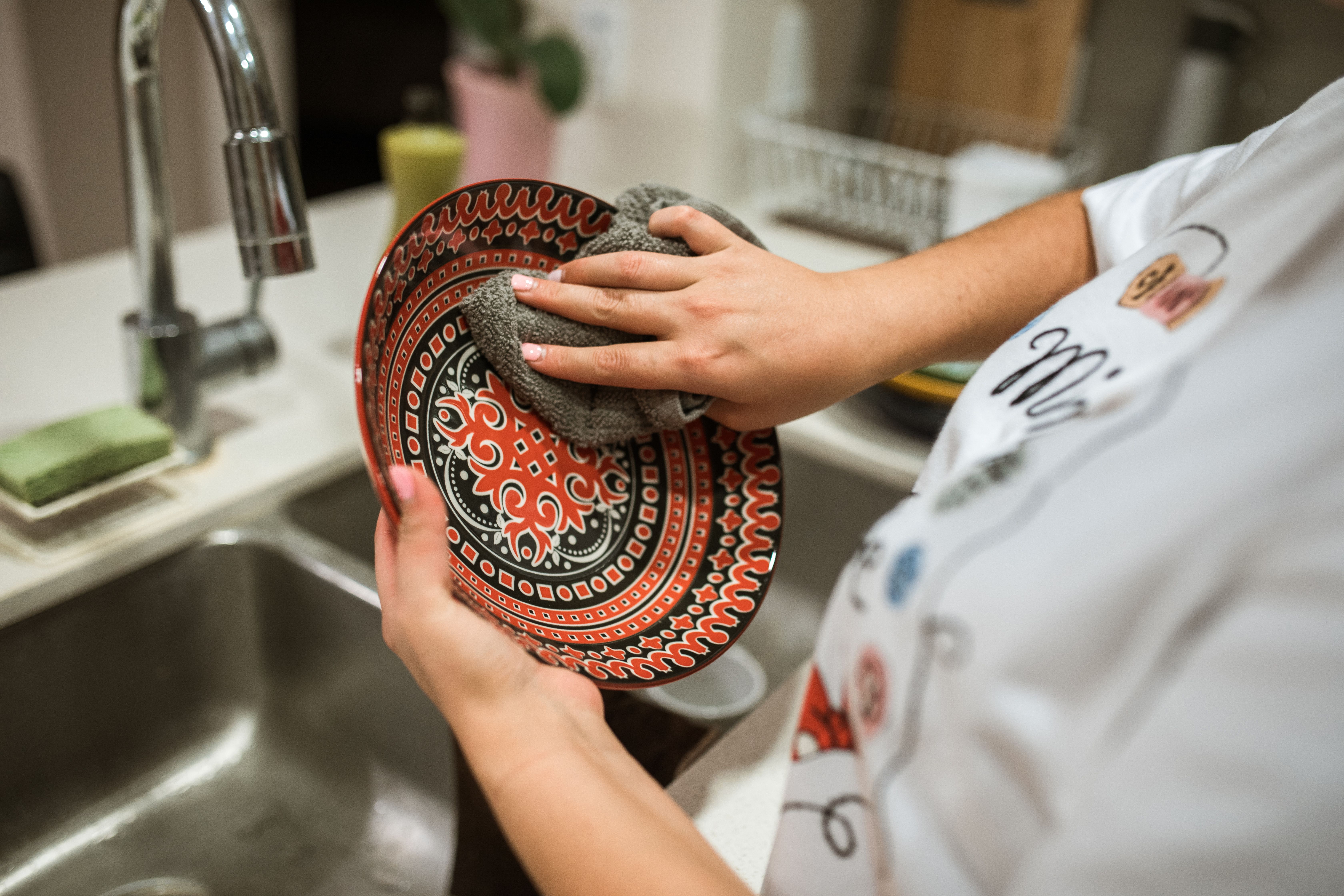 Pessoa com unhas cor-de-rosa lavando um prato vermelho e preto com desenhos circulares, usando uma esponja cinza sobre uma pia de cozinha de aço inoxidável