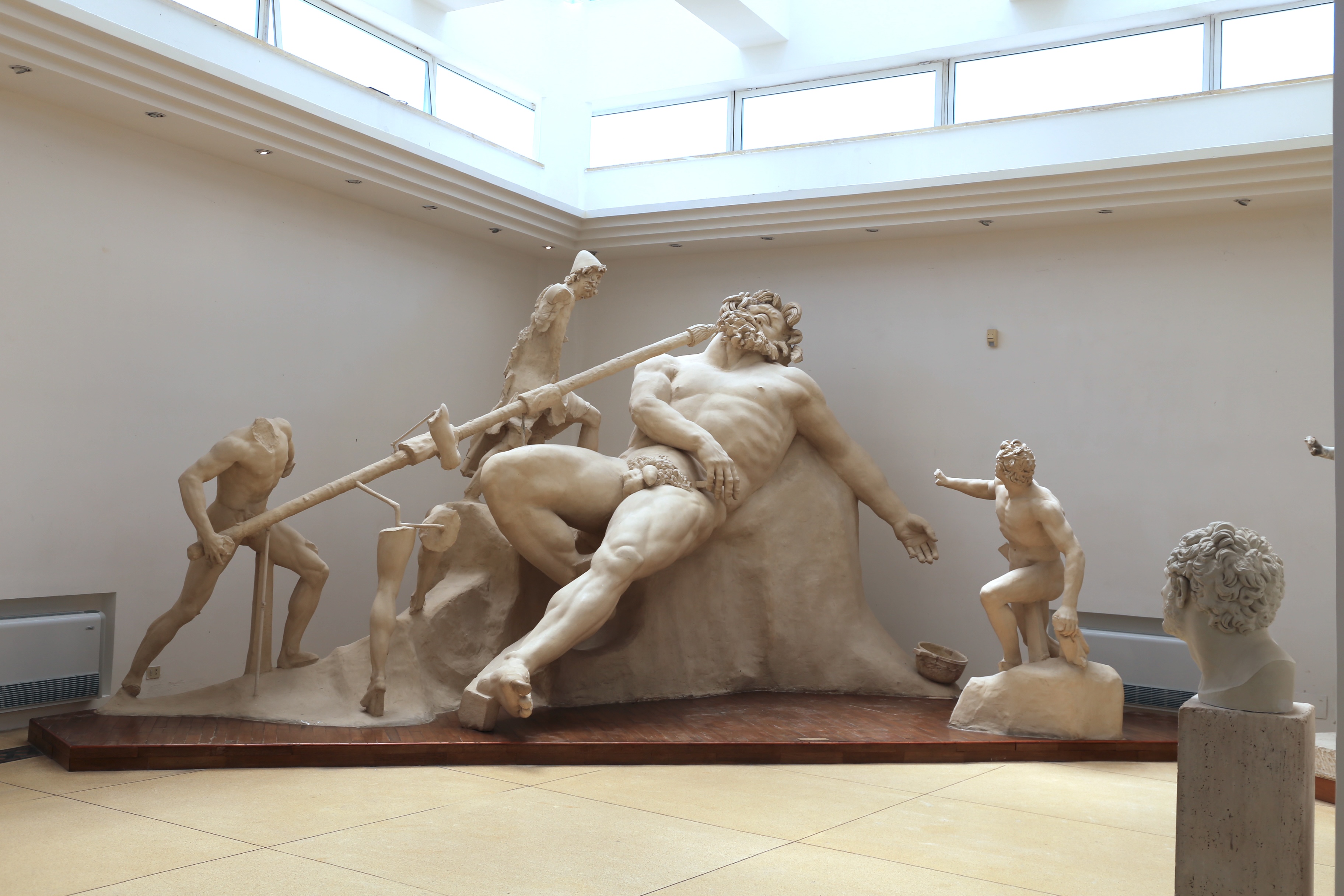 Escultura de gesso branco retratando Odisseu e seus homens cegando o ciclope Polifemo. Odisseu, um gigante barbudo, está deitado, com um tronco sendo empurrado em seu olho por três homens. Um quarto homem, à direita, observa a cena. Um busto de homem com cabelo encaracolado está em primeiro plano, à direita. O cenário é uma sala de museu com paredes claras e janelas no alto